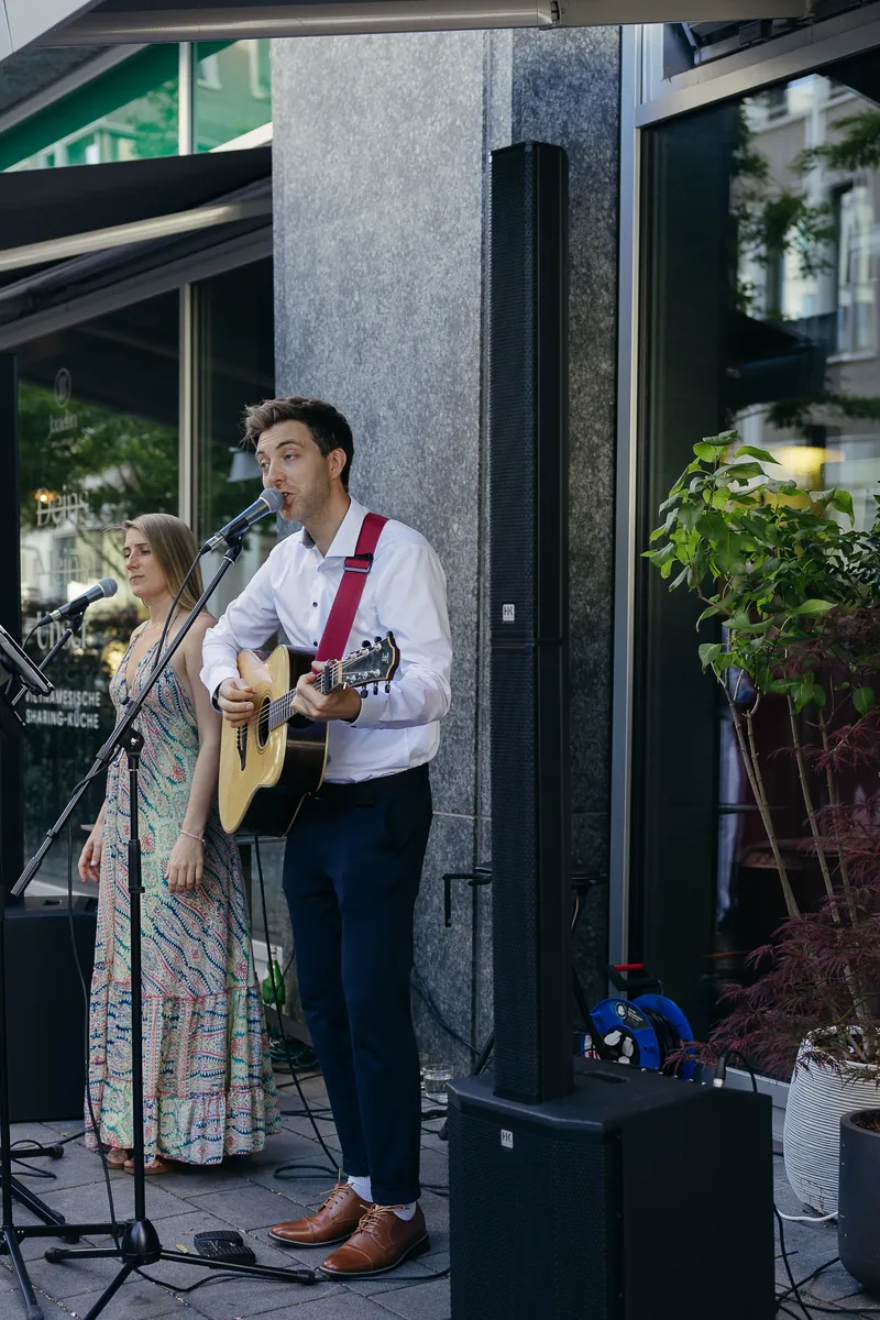 Jakob singt mit Gitarre, Samira im Hintergrund, Auftritt vor einem Restaurant in München